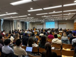 A group of about 50 conference-goers sit in chairs watching a presentation.