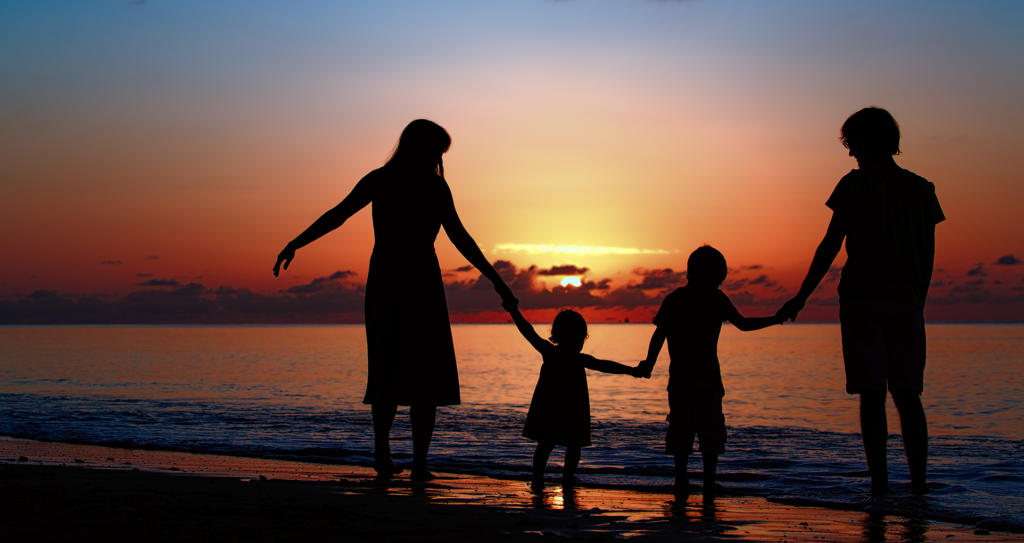 A family of four holding hands on the beach, silhouetted against the sunset.